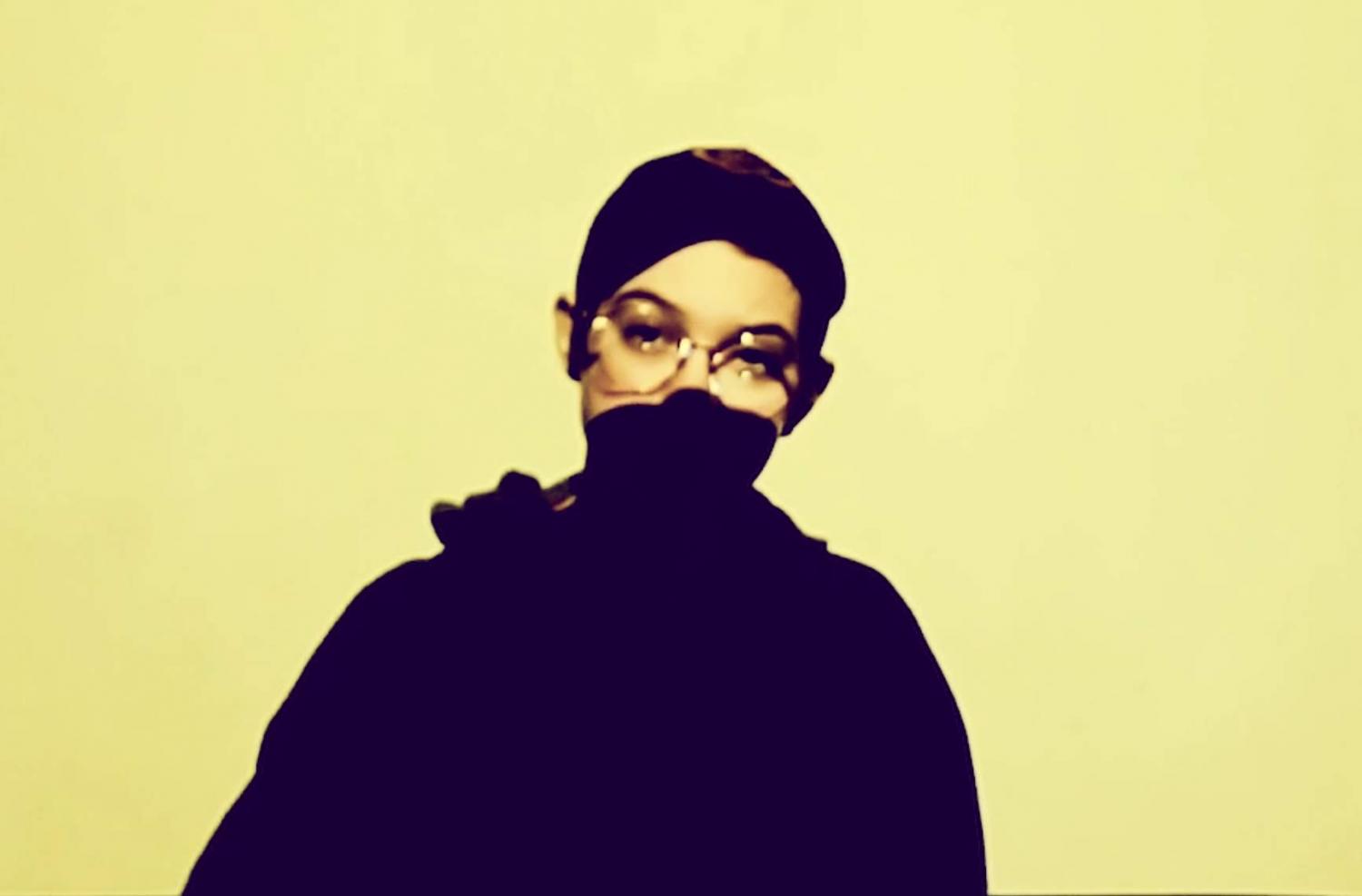 [Img description: A photo of Cameron standing in front of a blank background. He is wearing a black baseball cap and hoodie, with gold-rimmed glasses. A projector is casting yellow light at the wall, and shadow over the lower-half of his face.]