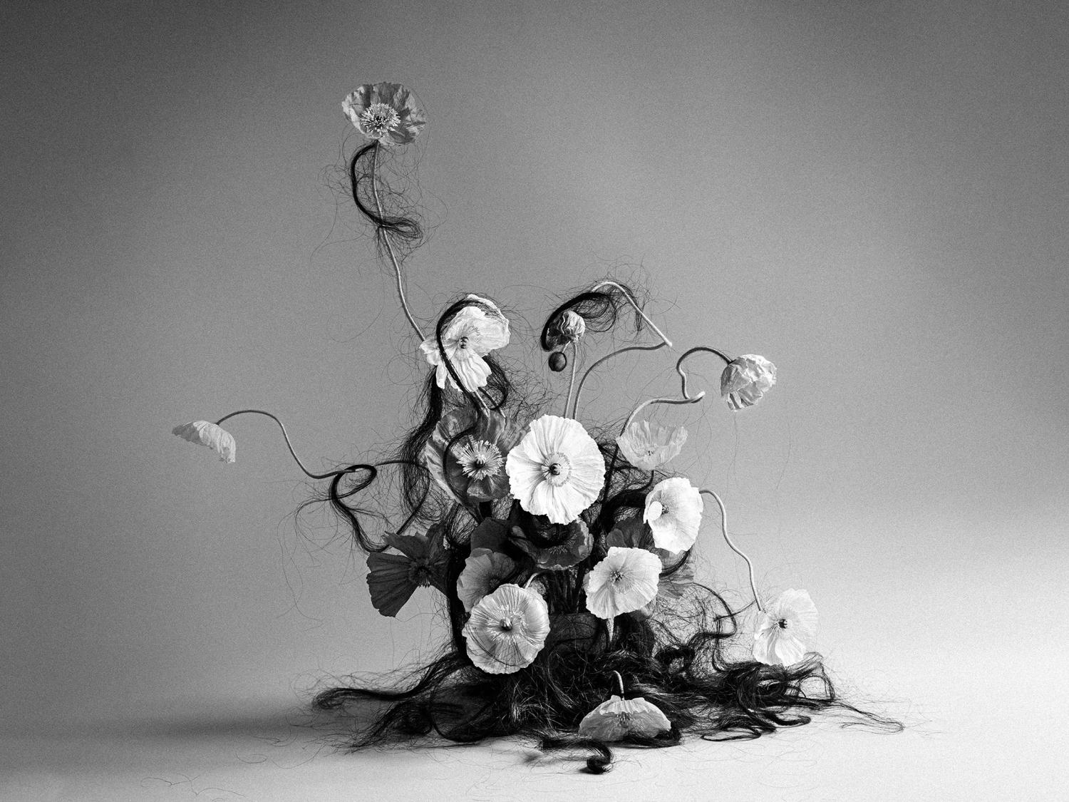 “Conversations with My Hair,” 2023, photograph. A black and white photograph of a group of poppies with dark human hair tangled around them.
