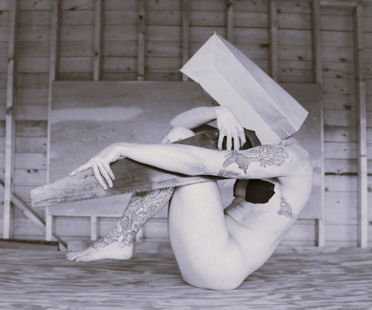 Image is ivan's piece "Won't Let Go". A black and white photo of a white person curled in a ball, with their toes off the ground, balancing on their seat/bum. They have a brown paper bag on their head and are curled over a hand saw, that is positioned parallel to the ground, perpendicular to their thigh in the position they are in.