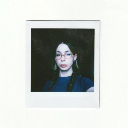 A digital scan of a dimly lit polaroid photograph. Pictured in the portrait is Brody, who appears in the centre wearing yellow-orange classes, a grey t-shirt, and her hair middle parted in a double braid. The background is dark. 