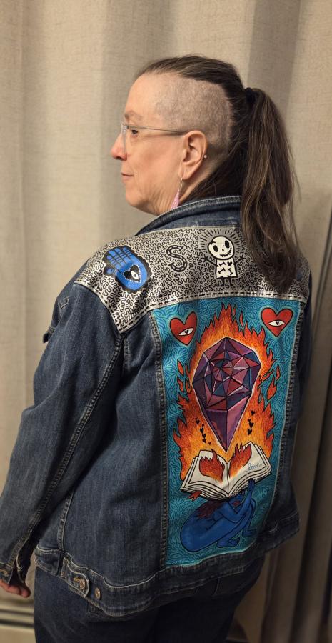 ID: Picture of Sheila, who is an older woman, with a mohawk looking over their shoulder while wearing a painted jean jacket. The jacket features an image of a large purple crystal above a flaming book held upon the back of a blue skinned woman. The jacket was painted by artist’s sister, Fiona Smyth.