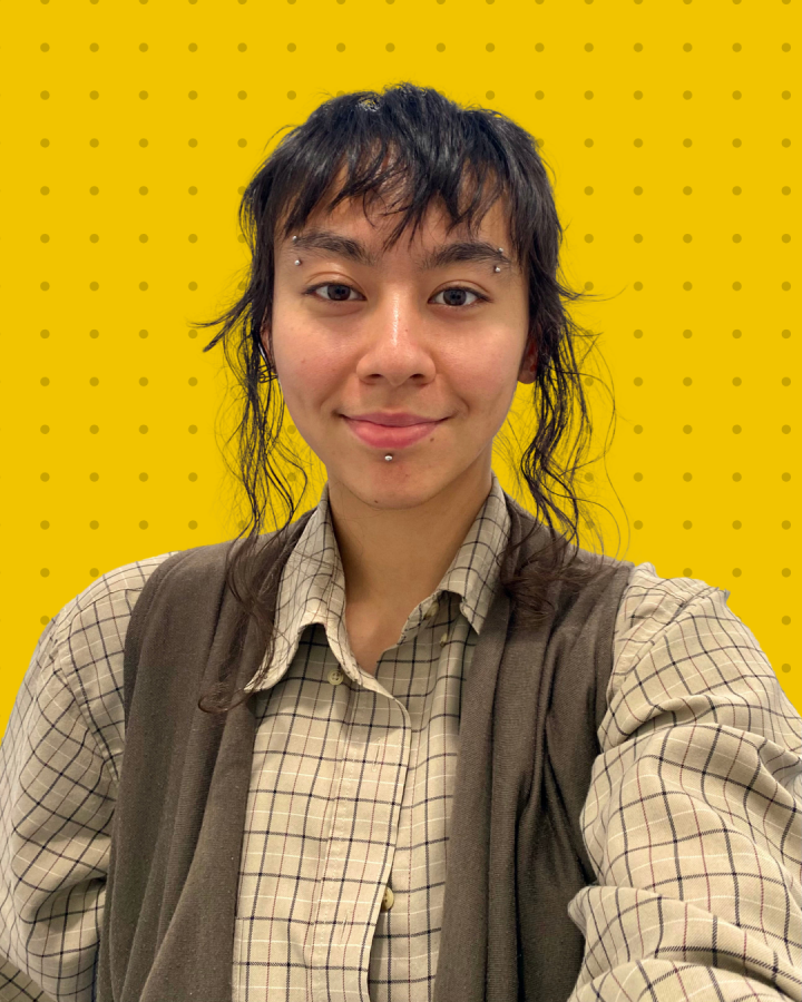 ID: Headshot of Ezra smiling and looking into the camera. They have medium-length dark brown hair, eyebrow and labret piercings, and are wearing a beige flannel top and green-grey vest. They are positioned against a yellow background with small, light polka dots.